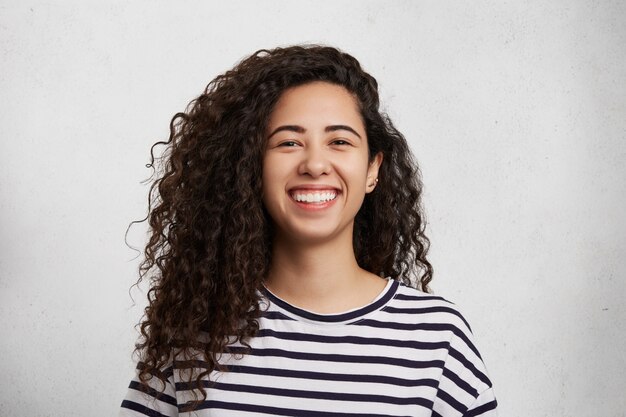 Portriat of pretty curly female wears striped black and whitte t shirt, smiles happily as being glad