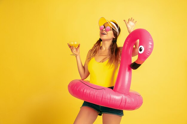Posing in beach ring with cocktail. Caucasian woman's portrait on yellow background. Beautiful female model in cap. Concept of human emotions, facial expression, sales, ad. Summertime, travel, resort.