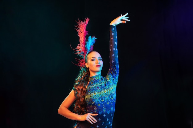 Posing. Beautiful young woman in carnival, stylish masquerade costume with feathers on black wall in neon light. Copyspace for ad. Holidays celebration, dancing, fashion. Festive time, party.