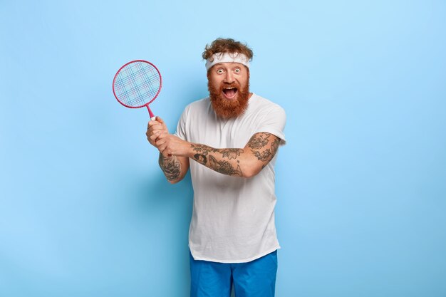 Positive active man plays tennis or badminton, has funny joyous expression, holds racket in hands, wears white headband