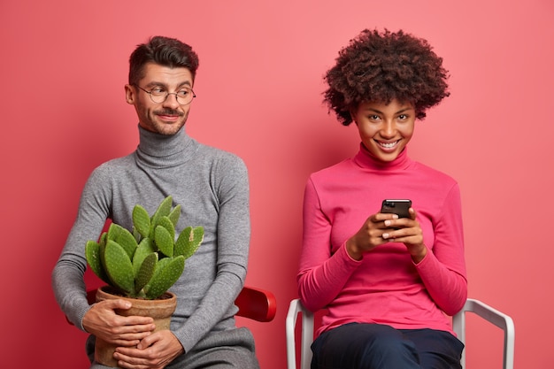 Positive addicted young woman uses mobile phone for browsing internet her boyfriend sits near holds potted cactus and needs live communication. Technology addiction and relationship concept.