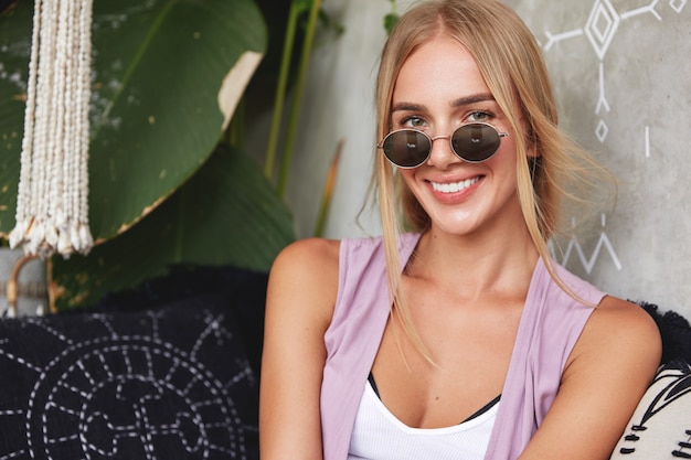 Positive adorable woman dressed casually, wears stylish sunglasses, sits on comfortable couch, waits for her order in restaurant, being in good mood after walk with boyfriend outdoor. Summer vacations