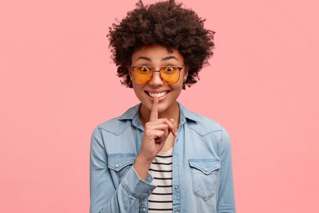 긍정적 인 아프리카 계 미국인 여성은 입술에 검지 손가락을 대고 비밀을 말하면서 자장 기호를 만들고 세련된 옷을 입고 행복한 표정을 짓고 분홍색에 고립 된 곱슬 머리와 하얀 치아를 보여줍니다.