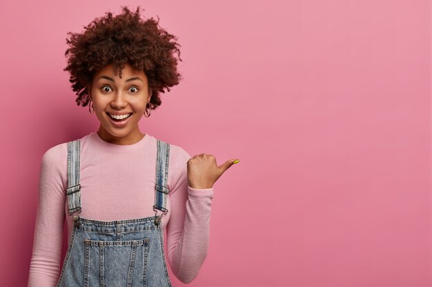 긍정적 인 아프리카 계 미국인 소녀가 엄지 손가락을 가리키고, 빈 분홍색 벽에 복사 공간을 보여주고, 행복하고 친근한 표현을하고, 캐주얼 한 옷을 입고, 실내 포즈를 취하고, 오른쪽으로 가라고 제안하고,이 방향을 따르라고 말합니다.