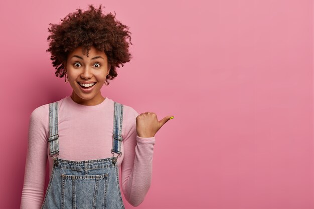 Positive African American girl points thumb, demonstrates copy space on blank pink wall, has happy friendly expression, dressed casually, poses indoor, suggests going right, says follow this direction