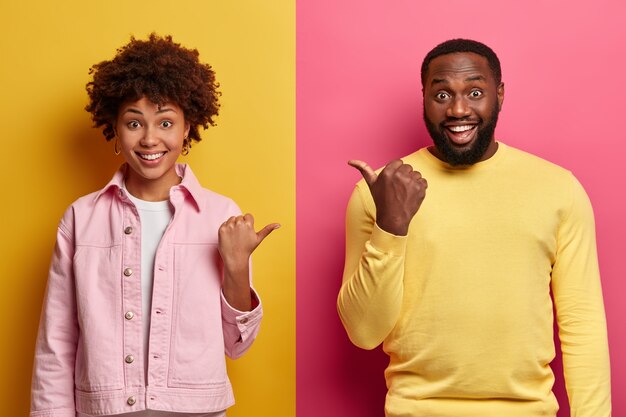 Positive African American woman and man point thumb at each other, smile and look enthusiastic, dressed in casual clothes, isolated on pink and yellow wall, have happy mood, friendly relations