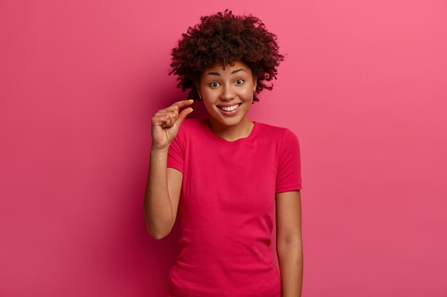 긍정적 인 아프리카 계 미국인 여성은 매우 작거나 작은 것을 보여주고, 매장에서 가격을 논의하고, 행복하게 미소를 짓고, 캐주얼 한 옷을 입고, 분홍색 벽에 고립 된 채 조금 말한다. 크기 개념.
