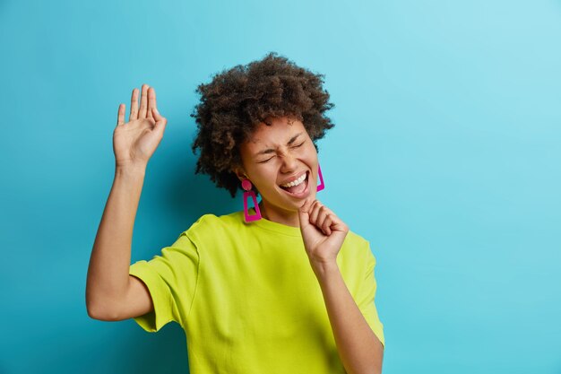 Positive African American woman sings song keeps hand as if microphone has fun has happy mood