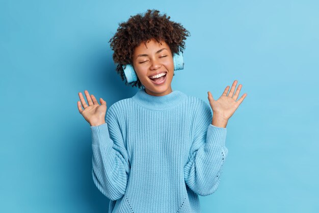 Позитивная афроамериканка с вьющимися волосами, поднимающая ладони, веселится во время прослушивания аудиозаписи в беспроводных наушниках, одетая в повседневный джемпер