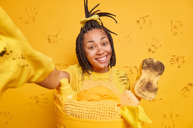 무료 사진 이빨 미소 더러운 얼굴을 가진 긍정적 인 아프리카 계 미국인 여자는 집에서 세탁을하는 동안 팔을 뻗어 자신의 사진을 만듭니다 흙은 노란색 벽에 고립 된 주말 동안 봄 청소를합니다