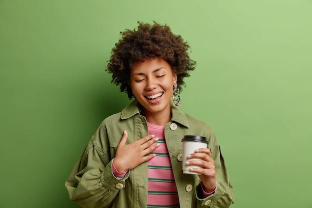 긍정적 인 아프리카 계 미국인 여자 eith 곱슬 머리는 행복에서 눈을 감고 심장 음료 테이크 아웃 커피 근처에 손을 유지 녹색 벽에 고립 된 유행의 옷을 입고 여가 시간을 즐긴다