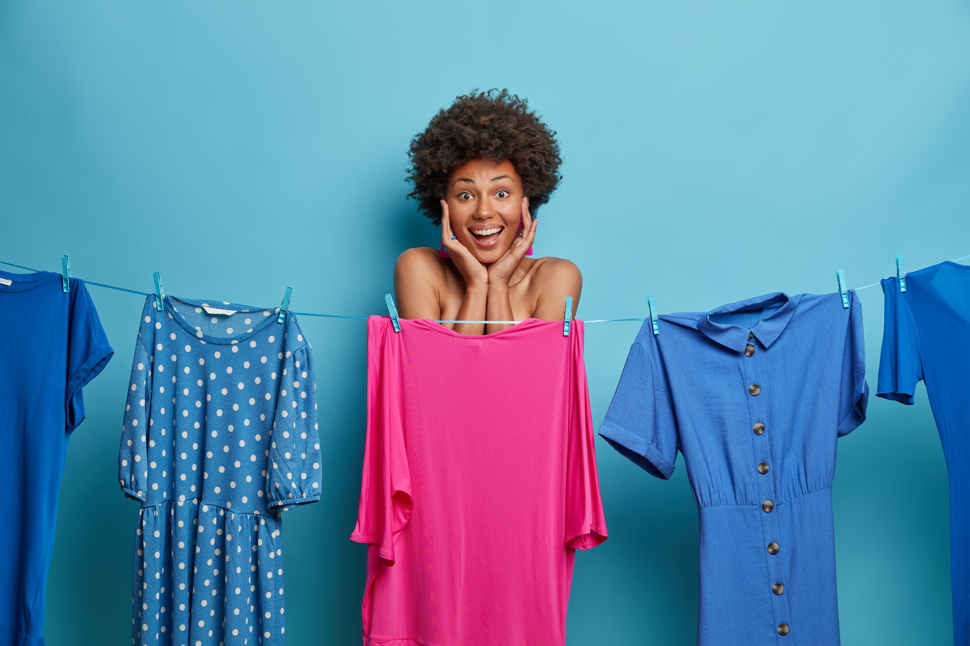 Positive Afro American woman keeps hands on face, hides naked body behind pink dress on rope, thinks what to wear for date, isolated on blue wall. Fashion, style, dressing and women concept