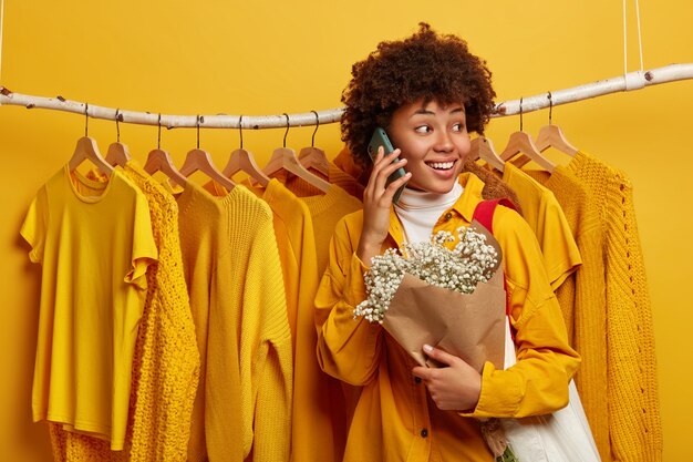 Positive Afro American woman turns from camera aside, has cheerful expression, stands against clothing rail, has phone talk