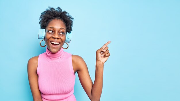 Positive Afro American woman with curly hair indicates at blank space