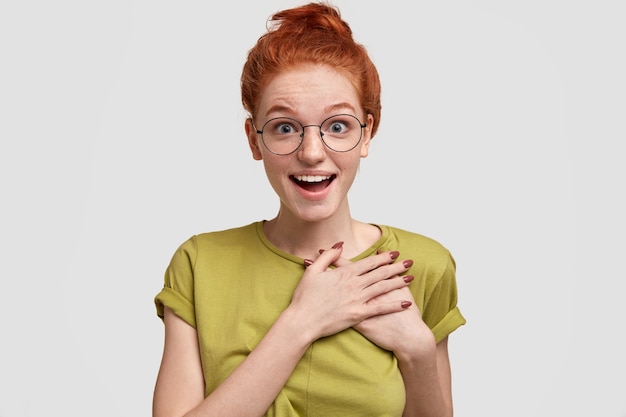 Positive amazed freckled red haired woman feels astonished, keeps hands on chest, wears round spectacles, stands against white wall