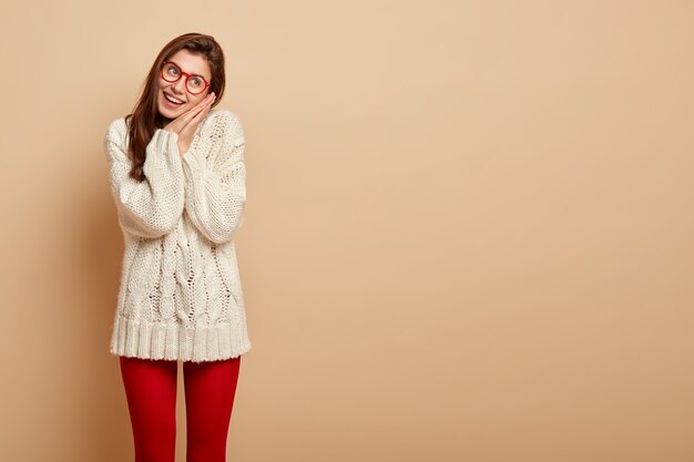 Foto gratuita la donna attraente e positiva inclina la testa di lato, tiene i palmi premuti insieme vicino al viso, ha un'espressione sognante