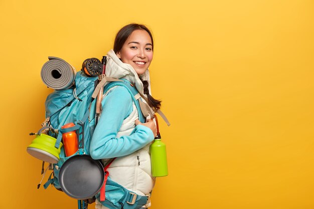 Positive Asian female hiker stands sideways to camera, carries big rucksack with necessary things for travel, has exciting adventurous trip, isolated over yellow wall