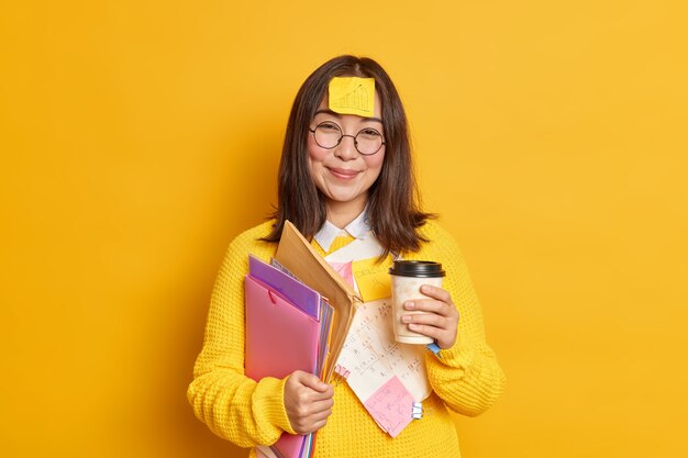 Positive Asian female worker holds disposable cup of coffee holds folders has sticker with graphic stuck on forehead has break after exam learning.