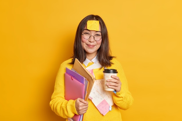 긍정적 인 아시아 여성 노동자는 일회용 컵 커피를 보유하고 있습니다. 폴더에는 이마에 그래픽이 붙어있는 스티커가 있습니다. 시험 학습 후 휴식이 있습니다.