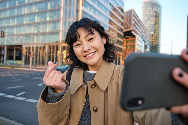긍정적인 아시아 소녀는 스마트폰으로 셀카를 찍고 손가락으로 마음을 보여주고 거리에서 사진을 찍습니다.