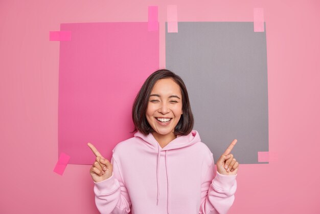 Positive Asian hipster girl wears casual hoodie suggests to check out link or banner indicates index finger at blank copy space