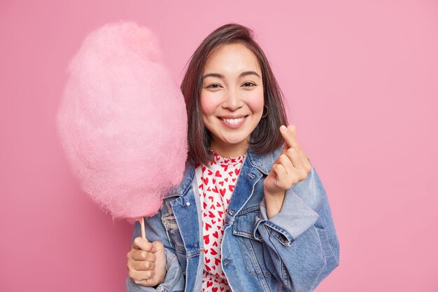 Positive Asian woman makes korean like sign expresses love smiles pleasantly expresses love dressed in denim jacket holds cotton candy on stick shapes mini heart isolated over pink wall