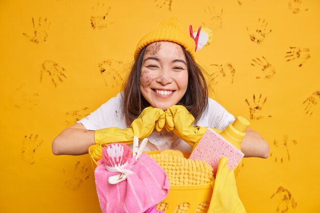Позитивная азиатская женщина улыбается, носит шляпу, защитные резиновые перчатки позирует возле корзины для белья, изолированной над желтой стеной