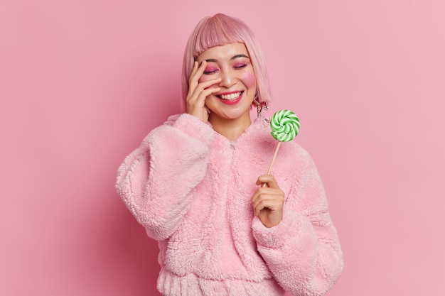 La donna asiatica positiva con i capelli rosa ha un trucco luminoso che tiene deliziose caramelle sul bastone