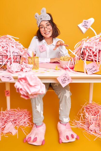 positive asian woman works from home during quarantine or lockdown dressed in nightwear prepares course work surrounded by cut paper poses at messy room against yellow wall
