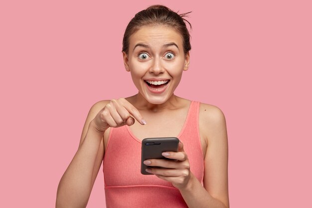 Positive astonished young female points at screen of mobile phone