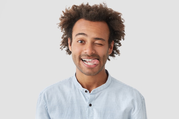 Positive attractive African American male with positive expression, shows tongue, has happy expression, stands against white wall, has crisp hair