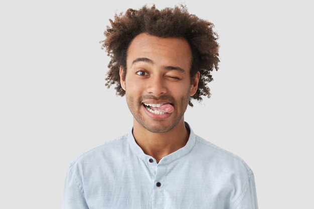 Позитивный, привлекательный афроамериканский мужчина с позитивным выражением лица, показывает язык, имеет счастливое выражение, стоит у белой стены, у него свежие волосы