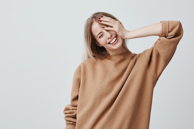 Positive attractive blonde young model wears casual loose sweater, being happy to receive good news. Joyful woman rejoices weekends, relaxing indoors