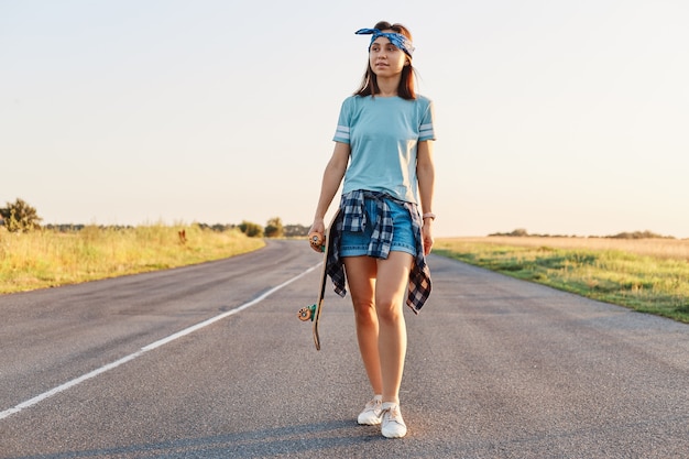 スケートボードを手に持って目をそらし、夏の日没のアスファルト道路を歩いて、短いTシャツとヘアバンドを身に着けているポジティブな魅力的な女性。