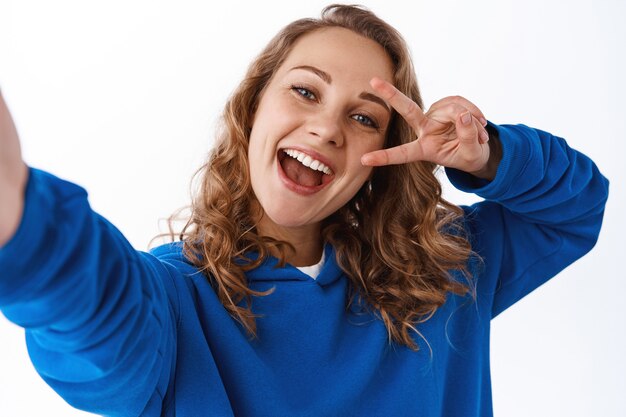 Positive attractive girl taking selfie, showing v-sign peace gesture near eye and holding camera with stretched hand, making photo on smartphone, white wall