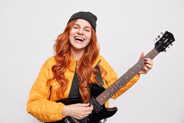 Positive attractive teenage girl talented popular singer plays acoustic guitar presents her new rock song has long ginger hair wears hat orange jacket 