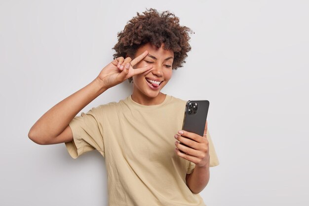 Positive attractive woman with curly hair makes peace gesture over eye sticks out tongue poses for selfie in smartphone screen dressed in casual beige t shirt isolated over white background