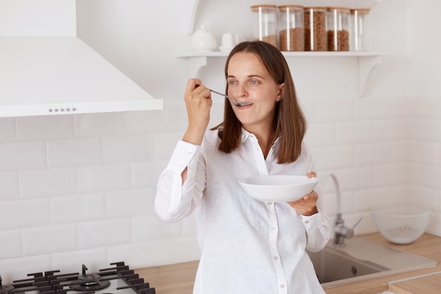 Positive attractive young adult woman with dark hair wearing white casual shirt. having breakfast, holding plate in hands, looking at away, eating from spoon.
