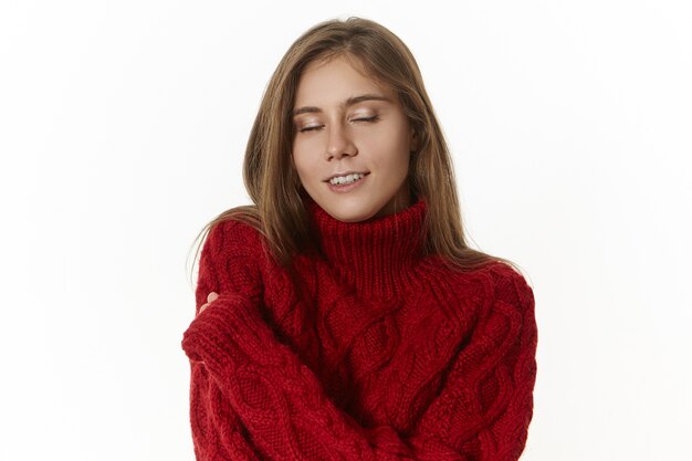 positive attractive young Caucasian woman with long loose hair closing eyes with pleasure, keeping arms around shoulders, enjoying warm cozy knitted sweater, smiling joyfully