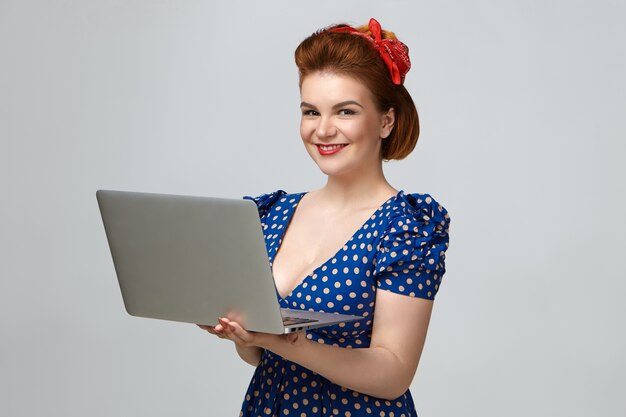 Positive attractive young lady wearing vintage dress and red lipstick posing in studio, enjoying high speed wireless internet connection using portable computer. People