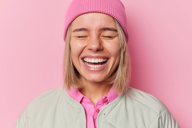 Positive authentic European woman smiles broadly shows white teeth giggles happily with closed eyes wears hat and jacket poses in studio against pink background People and emotions concept