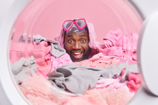 Positive bearded Afro American man covered with laundry has fun while washing dirty clothing at home poses from inside of washing machine wears snorkeling mask on forehead