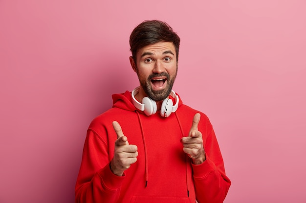 Positive bearded hipster guy points directly at you, makes good choice, says excellent, makes its up to you gesture, wears red hoodie and modern headphones, poses against pink pastel wall