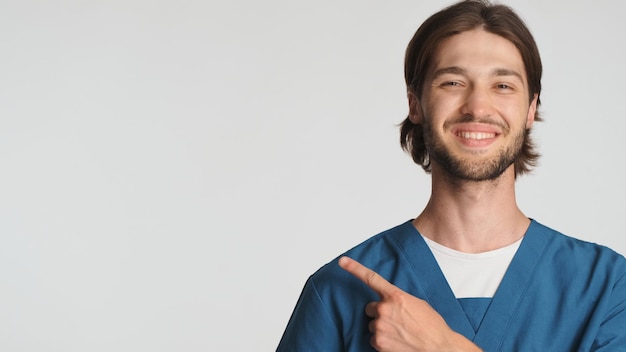 Positive bearded male doctor smiling points away on space for text over white background Attractive intern wearing uniform looking confident isolated
