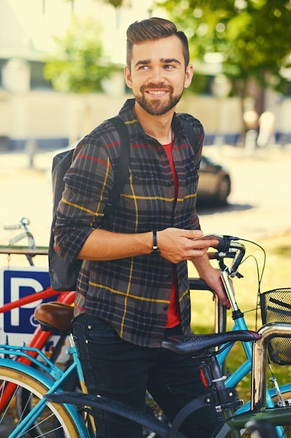 양털 셔츠를 입은 긍정적인 수염 남성이 자전거 주차장 근처에서 스마트폰을 사용하고 있습니다.