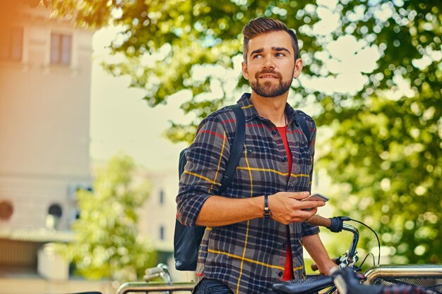 A positive bearded male dressed in a fleece shirt using a smart phone near bicycle parking.