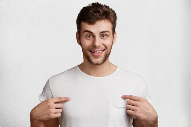 Positive bearded male with stubble, blue eyes and satisfied expression, indicates at blank copy space on t shirt, isolated over white background. People, good feelings and advertisment concept