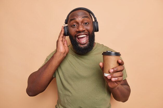 Positive bearded man has fun listens music via headphones drinks takeaway coffee