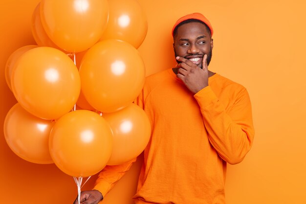 Positive bearded man holds bunch of balloons celebrates festive occasion wears casual jumper and hat isolated over orange wall being on party