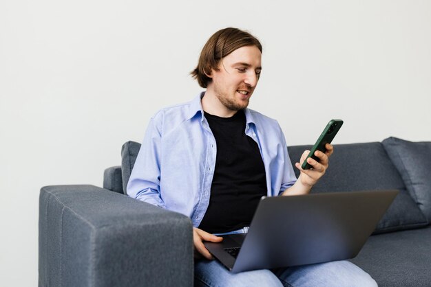 Позитивный бородатый мужчина с ноутбуком разговаривает по смартфону, сидя на диване в гостиной